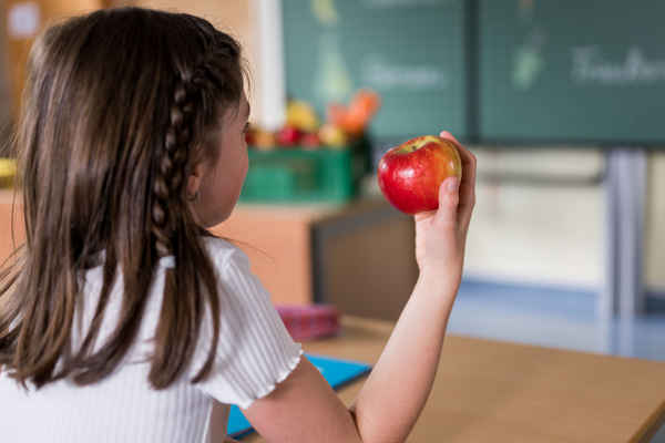 KI generiert: Mädchen im Klassenzimmer hält einen Apfel in der Hand. Im Hintergrund eine Tafel und Obstkiste.