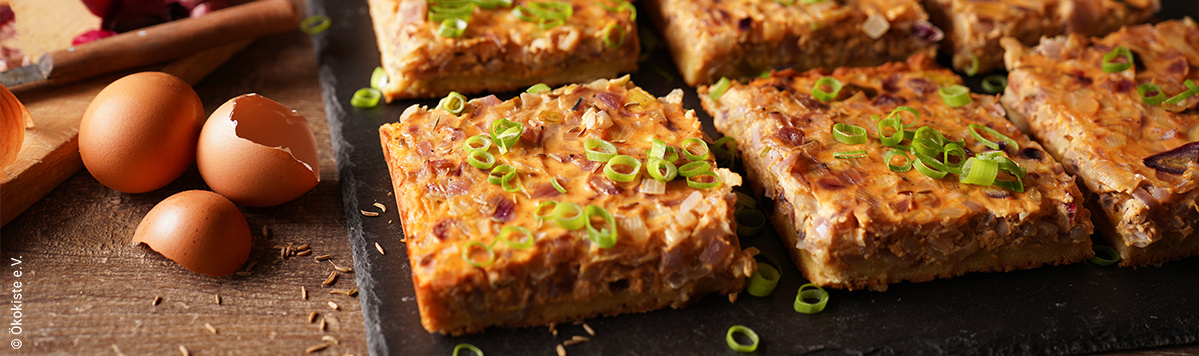 KI generiert: Ein Zwiebelkuchen in Stücken, garniert mit Frühlingszwiebeln. Links leere Eierschalen.© Ökokiste e.V.