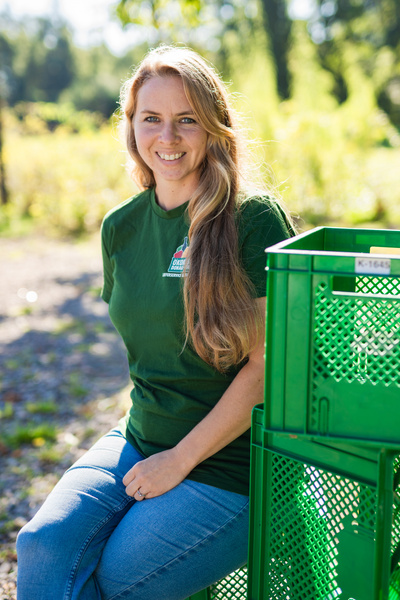 KI generiert: Eine Person mit langem Haar sitzt draußen neben grünen Kisten, lächelt und trägt ein grünes Shirt.