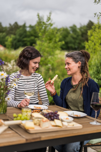 KI generiert: Zwei Frauen sitzen an einem Tisch im Freien und unterhalten sich, umgeben von Käse und Wein.