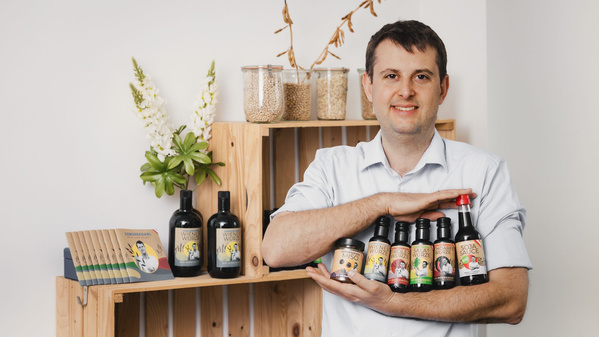 KI generiert: Ein Mann steht lächelnd vor einem Regal mit verschiedenen Flaschen und Produkten, während er weitere Flaschen in seinen Armen hält. Das Bild vermittelt den Eindruck eines Geschäfts oder einer Produktpräsentation.