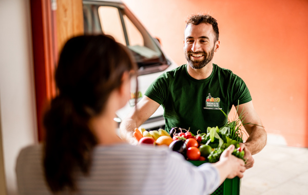 KI generiert: Ein Mann liefert eine Kiste mit frischem Gemüse an eine Frau. Auf dem Shirt steht: "Ökokiste".
