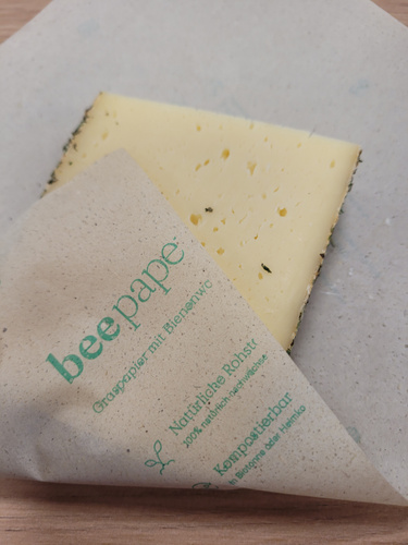 KI generiert: Käse in umweltfreundlichem Papier eingewickelt. Text: „beepape, Graspapier mit Bienenwachs, Natürliche Rohst…“.