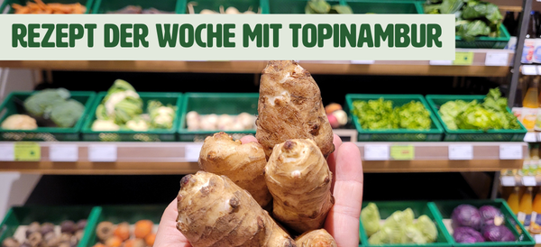KI generiert: Das Bild zeigt eine Hand, die Topinambur-Knollen hält, vor einem Gemüseregal in einem Supermarkt. Darüber steht der Text "Rezept der Woche mit Topinambur".