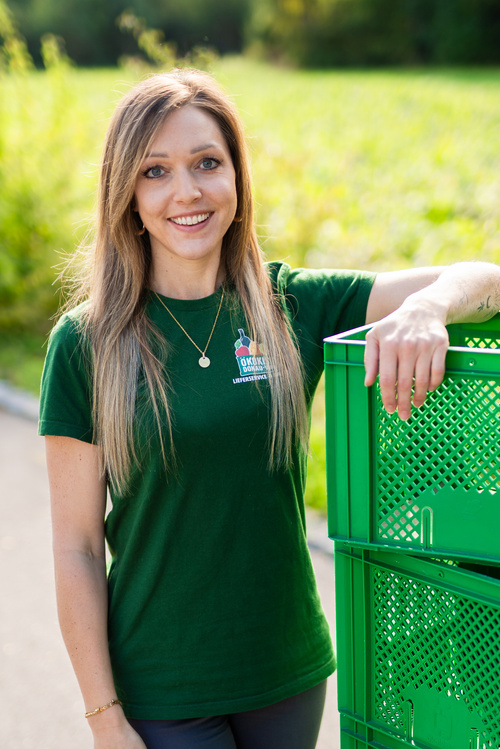 KI generiert: Eine Frau steht draußen, lehnt an einer grünen Kiste und trägt ein T-Shirt der "Ökokiste Bamberg".