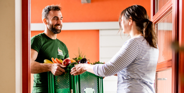 KI generiert: Ein Mann liefert einer Frau eine Kiste mit frischem Obst und Gemüse an der Tür.