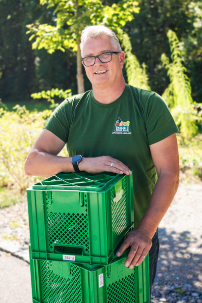 KI generiert: Ein Mann mit Brille steht draußen neben grünen Kisten. Text: "Ökokiste - Überzeugung & Qualifikation".