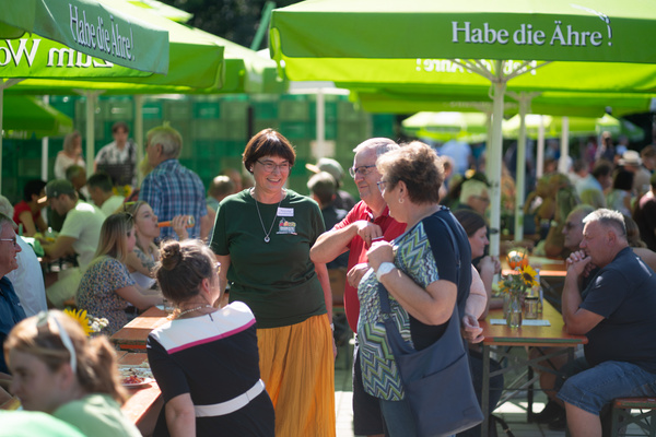 KI generiert: Eine Gruppe von Leuten plaudert auf einem sonnigen Außenfest. Text: "Habe die Ähre!" auf grünen Schirmen.