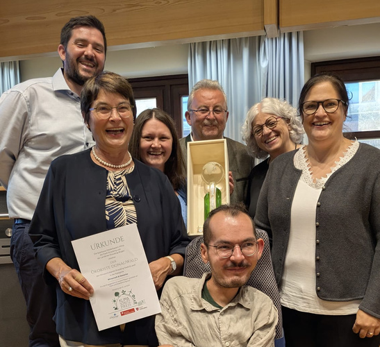 KI generiert: Eine Gruppe von Menschen hält eine Urkunde mit dem Text "ÖKOKISTE DONAUKWALD".