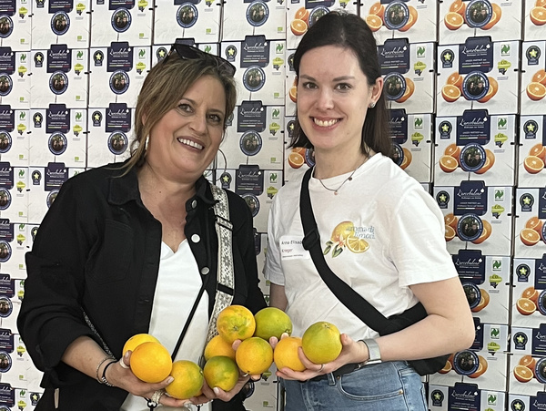 KI generiert: Zwei Personen halten Orangen vor einer Wand mit Orangenboxen. Text auf Shirt: "Arance di Zagara" mit Orangenmotiv.