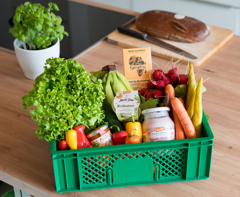 KI generiert: Eine grüne Kiste voller frischem Obst, Gemüse und Produkten in einer Küche. Text: "fairafric", "Kräuter".