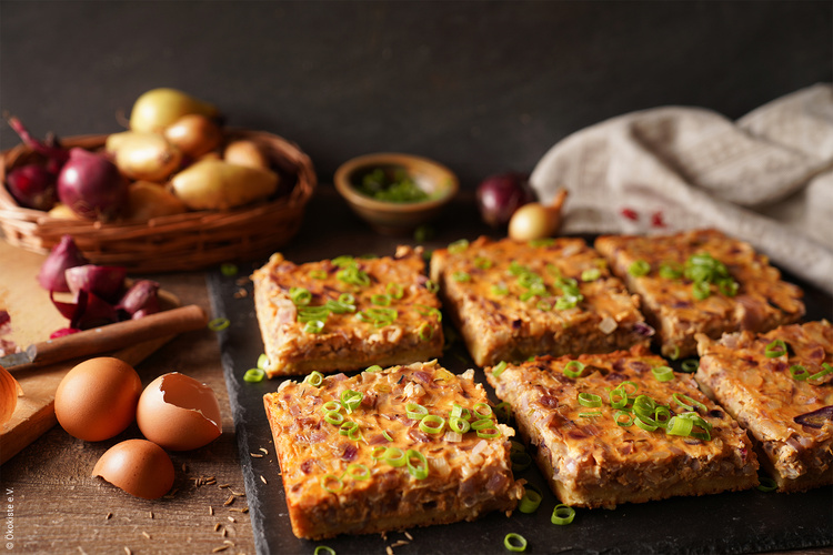 KI generiert: Zwiebelkuchenstücke mit Frühlingszwiebeln garniert auf einem Brett, im Hintergrund Zwiebeln im Korb.