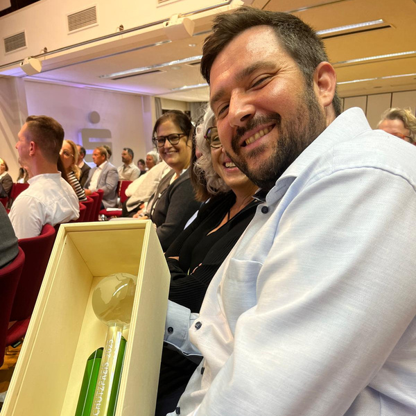 KI generiert: Ein lächelnder Mann hält eine Auszeichnung in einem Holzkasten, umgeben von anderen Personen im Publikum.