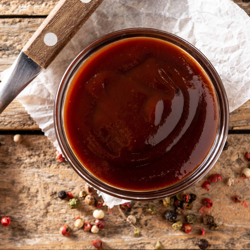KI generiert: Ein Glas Barbecue-Sauce auf Holzbrett mit Pfefferkörnern drumherum.