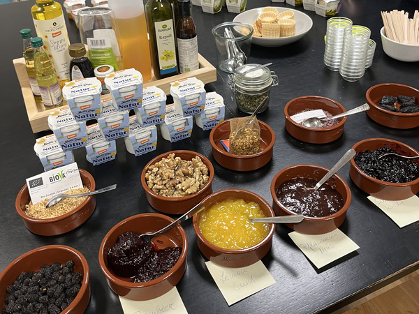 KI generiert: Eine Auswahl an Aufstrichen, Nüssen und Joghurt auf einem Tisch; Beschriftungen für Brombeere, Lemon Curd.