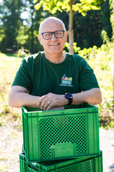 KI generiert: Eine Person lehnt lächelnd an grünen Kisten im Freien. Text: "Ökokiste Donauries".