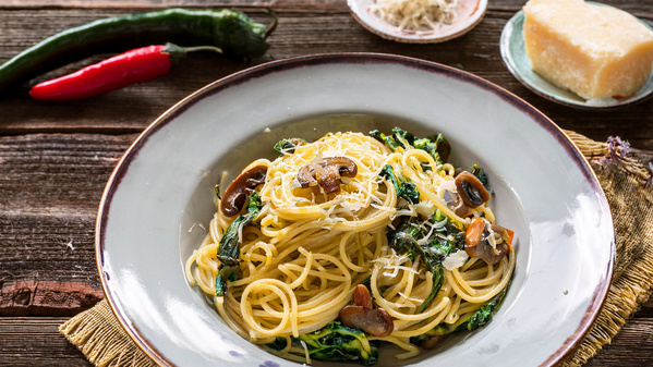KI generiert: Ein Teller Spaghetti mit Gemüse und Pilzen, umgeben von Chilis und Parmesan auf einem Holztisch.