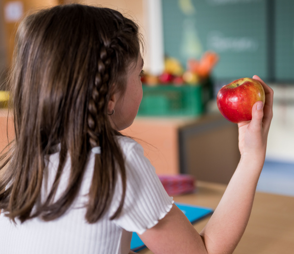 KI generiert: Ein Mädchen hält einen roten Apfel in einem Klassenzimmer. Im Hintergrund ist verschwommen eine Tafel zu sehen.