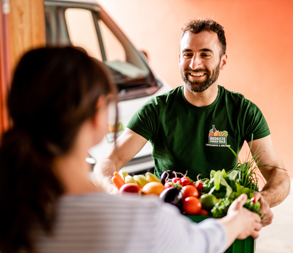 KI generiert: Ein Mann übergibt einer Frau eine Kiste voller Gemüse. Sein T-Shirt trägt das Logo "Ökokiste".