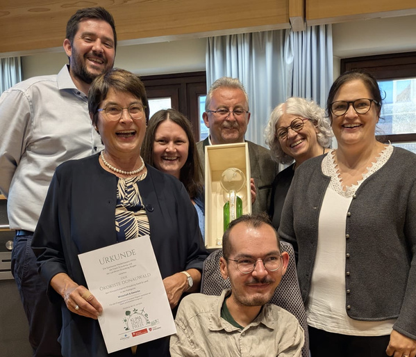 KI generiert: Eine Gruppe fröhlicher Menschen hält eine Urkunde und eine Trophäe. Text: "URKUNDE ÖKOKISTE DONAUWALD."