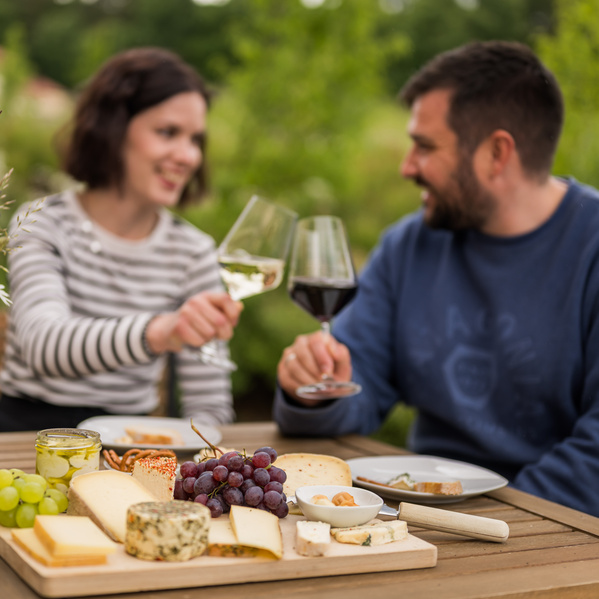 KI generiert: Ein Paar stoßt mit Wein an, vor einem Käsebrett mit Trauben im Freien.