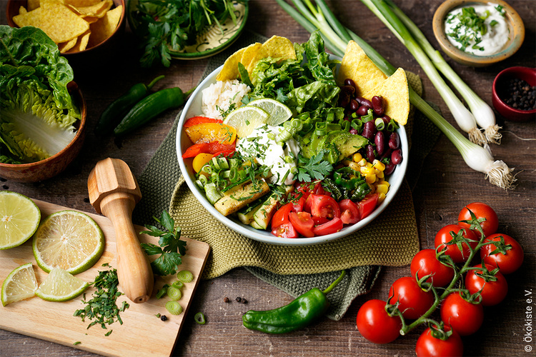 KI generiert: Ein bunter Salat mit Gemüse und Tortillachips, umgeben von frischem Gemüse. Text: "© Ökokiste e. V."