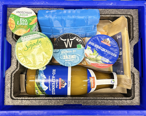 KI generiert: Eine Kühlbox mit Bio-Lebensmitteln, darunter Joghurt, Saft und Tzatziki. Text: "Bio Lassi", "Sojade", "ZAZIKI".