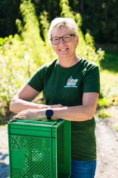 KI generiert: Eine Person mit Brille lehnt sich lächelnd auf eine grüne Kiste im Freien. Text: "DIE GEMÜSEKISTE".