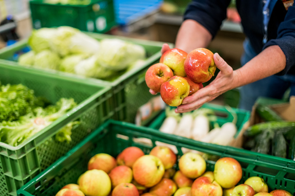 KI generiert: Person hält Äpfel in der Hand, umgeben von frischem Obst und Gemüse in grünen Kisten.