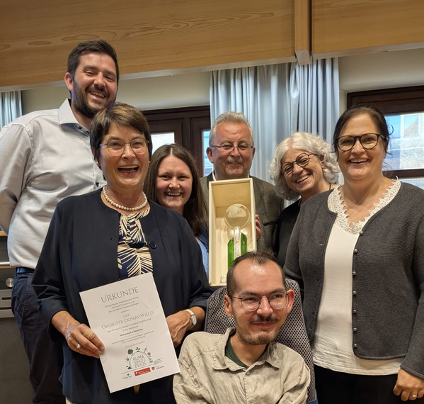 KI generiert: Eine Gruppe von Menschen hält eine Urkunde und eine Auszeichnung in die Kamera. Text: "Urkunde, Ökokiste Donauwald".