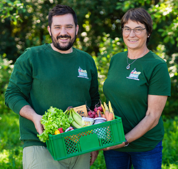 KI generiert: Das Bild zeigt zwei Personen hinter einem Tisch, der mit einer Vielzahl von frischem Obst und Gemüse bedeckt ist. Es gibt ein Werbeschild, das darauf hinweist, dass die sechste Kiste kostenlos ist.