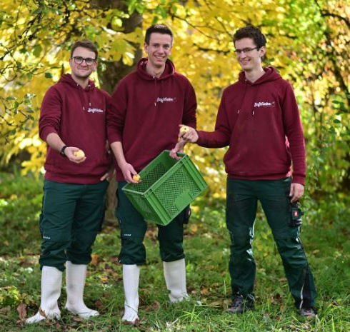 KI generiert: Drei Personen in roter Arbeitskleidung halten einen Korb und Äpfel in einem herbstlichen Garten.