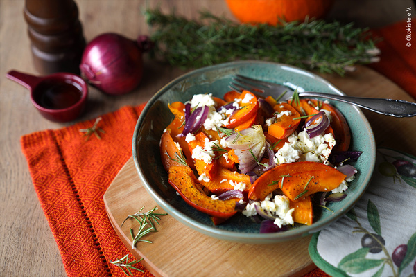 KI generiert: Das Bild zeigt einen Teller mit einem herbstlichen Salat aus Kürbis, roten Zwiebeln und Feta, garniert mit Rosmarin. Im Hintergrund sind verschiedene Zutaten und Gewürze zu sehen, darunter eine rote Zwiebel und ein Rosmarinzweig.