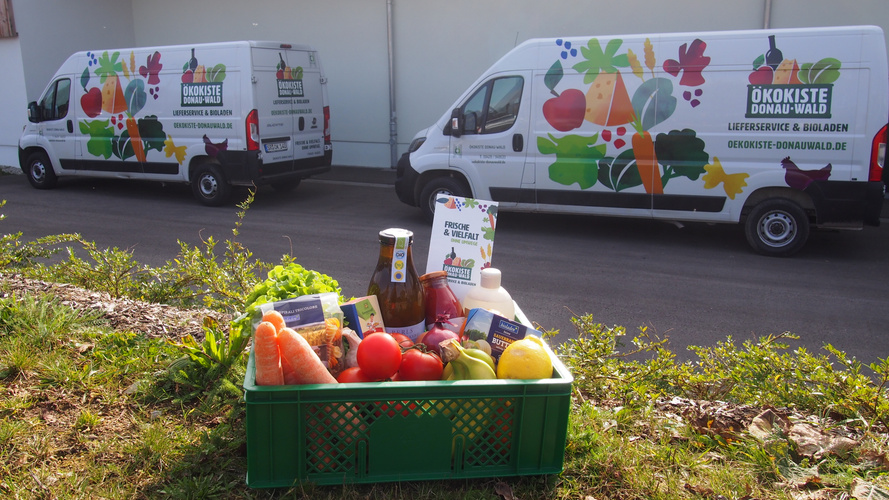 KI generiert: Ein Korb mit frischem Obst und Gemüse vor Lieferwagen. Text: "Ökokiste Donau-Wald", "FRISCHE & VIELFALT".