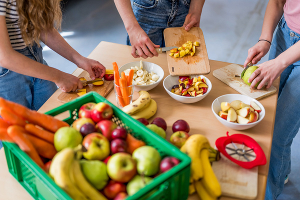 KI generiert: Drei Personen schneiden Obst und Gemüse in der Küche. Ein Korb mit frischem Obst steht auf dem Tisch.