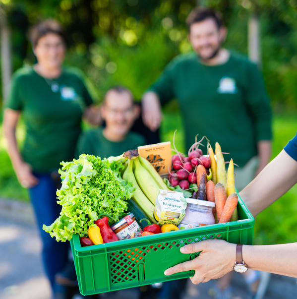 KI generiert: Ein Korb mit frischem Obst und Gemüse wird gehalten. Hintergrund unscharf mit Personen.