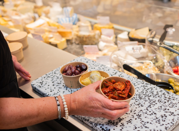 KI generiert: Das Bild zeigt eine Person, die in einem Feinkostladen vor einer Theke mit verschiedenen Käsesorten steht und eine Platte mit kleinen Schälchen von Oliven und getrockneten Tomaten hält. Der Fokus liegt auf den angebotenen Lebensmitteln auf der Theke und in den Schälchen.