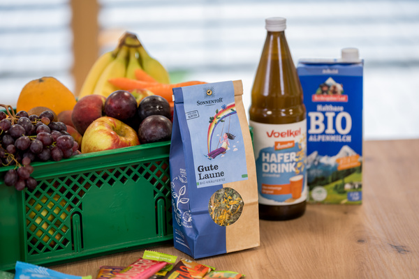 KI generiert: Korb mit Obst, Tee, Haferdrink und Biomilch auf einem Tisch. Text: „Gute Laune BIO KRÄUTERTEE“.