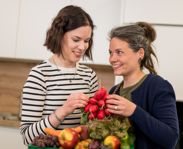KI generiert: Zwei Frauen halten frisches Obst und Gemüse in einem Raum.