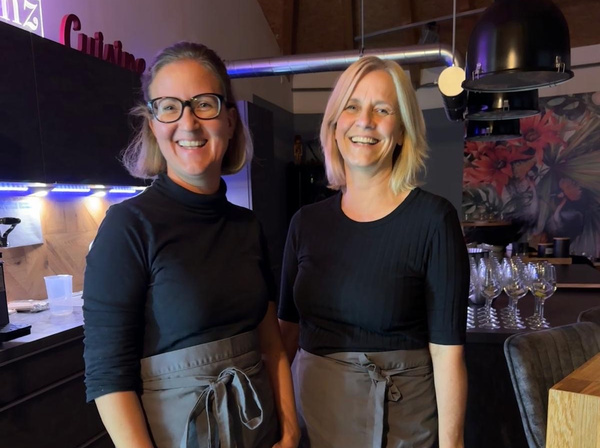 KI generiert: Zwei lächelnde Frauen in einer Küche, die beide Schürzen tragen. Im Hintergrund Gläser und Dekorationen.