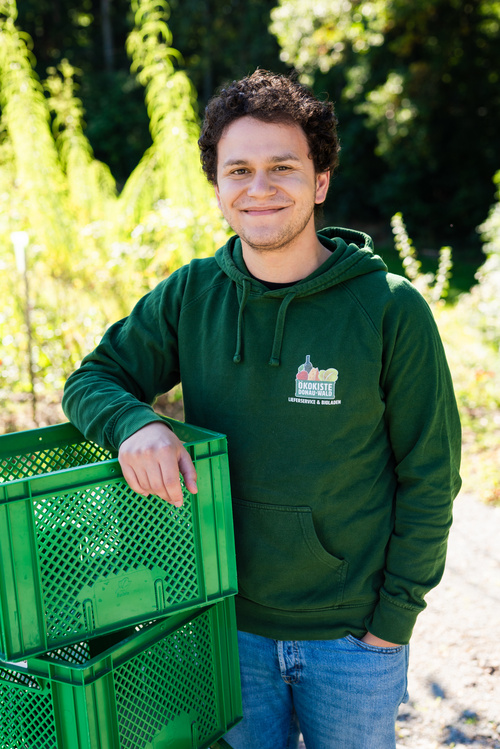 KI generiert: Eine Person in grüner Kleidung steht neben grünen Kisten im Freien. Text: "ÖKOKISTE".