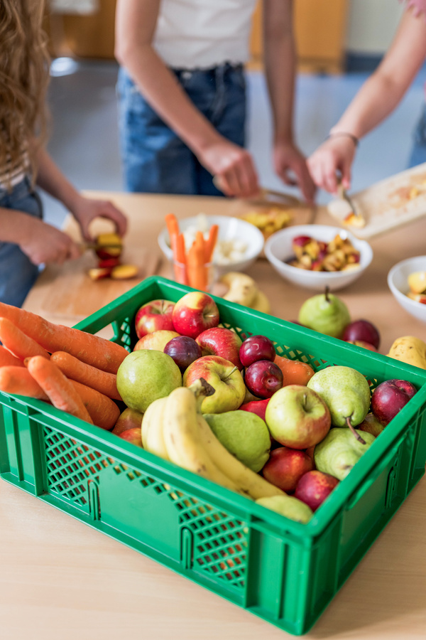 KI generiert: Kiste mit buntem Obst und Gemüse auf einem Tisch, Menschen schneiden und snacken im Hintergrund.