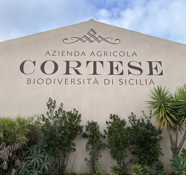 KI generiert: Gebäude mit der Aufschrift: "AZIENDA AGRICOLA CORTESE BIODIVERSITÀ DI SICILIA", umgeben von Pflanzen.