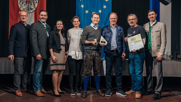 KI generiert: Eine Gruppe von acht Personen posiert für ein Foto, einige halten Auszeichnungen in den Händen. Im Hintergrund sind die Flaggen von Österreich, der Europäischen Union und einer Stadtflagge zu sehen.