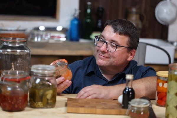 KI generiert: Ein Mann betrachtet ein Glas eingelegtes Gemüse in einer Küche mit verschiedenen Vorratsgläsern.
