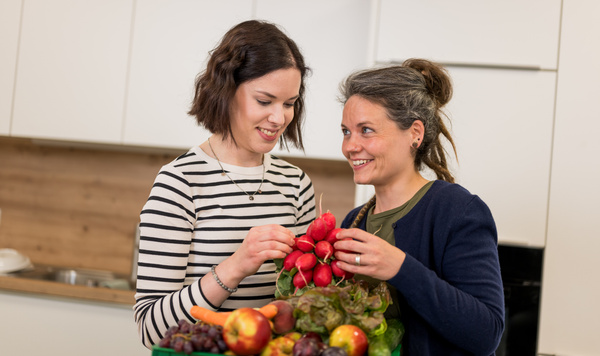 KI generiert: Zwei Frauen halten eine große Auswahl an frischem Obst und Gemüse in einer Küche.
