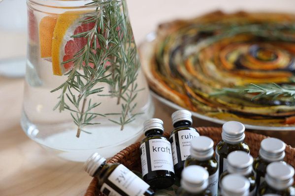 KI generiert: Ein Krug mit Rosmarin und Zitrusfrüchten, mehrere Fläschchen und ein buntes Gemüsegericht. Text: "kraft", "ruhe".