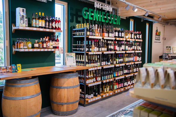 KI generiert: Das Bild zeigt einen Verkaufsbereich mit einem Regal voller Weinflaschen und Spirituosen sowie einem Tisch aus Holzfässern. An der Wand steht der Schriftzug "DAS LEBEN GENIESSEN".