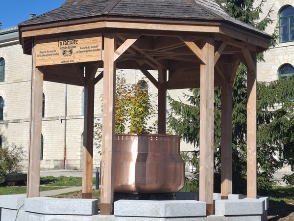 KI generiert: Ein Pavillon aus Holz mit einem großen, kupfernen Kessel. Text: "Juraflore - record du monde de la plus grosse fondue".
