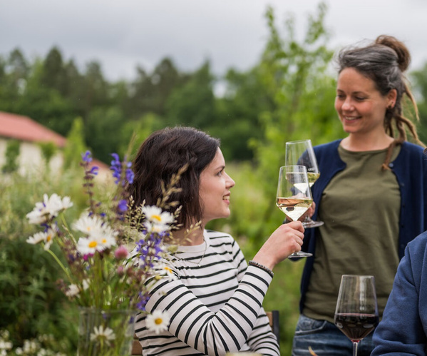 KI generiert: Zwei Personen stoßen mit Weingläsern an, Blumen im Vordergrund, grüne Natur im Hintergrund.